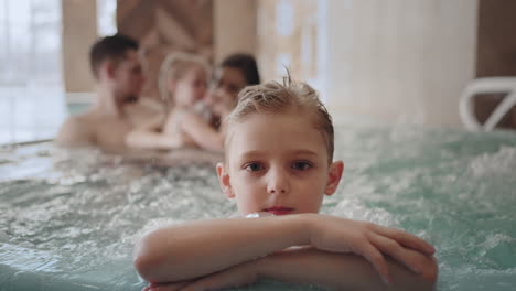 cute-little-boy-is-looking-at-camera-in-water-park-family-is-chilling-warm-bath-in-pool